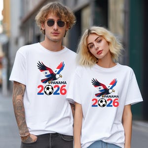 Pu&ograve; includere: Magliette bianche con una grafica di un'aquila con ali rosse, bianche e blu, la scritta "PANAMA", un pallone da calcio e il numero "2026". Le magliette sono indossate da due persone.