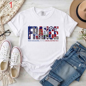 May include: White t-shirt with the word "FRANCE" in colorful letters, featuring iconic French symbols. Below the word is the phrase "NOS COULEURS, NOTRE COEUR, NOTRE FOI!" The shirt is paired with blue jeans and white sneakers.