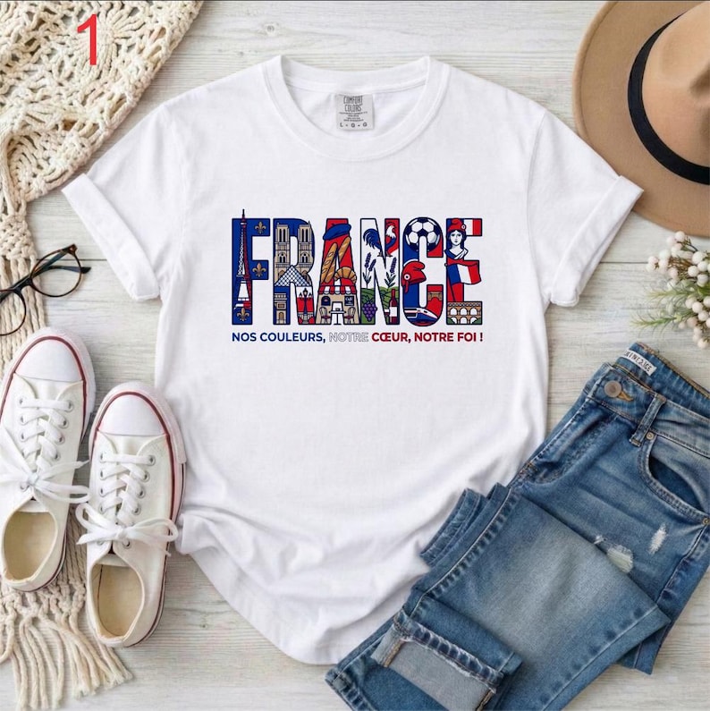 May include: White t-shirt with the word "FRANCE" in colorful letters, featuring iconic French symbols. Below the word is the phrase "NOS COULEURS, NOTRE COEUR, NOTRE FOI!" The shirt is paired with blue jeans and white sneakers.
