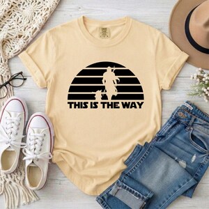 May include: Cream-colored t-shirt featuring a black graphic of a figure and a small companion against a striped background. The text "THIS IS THE WAY" is below the graphic. The shirt is paired with blue jeans and white sneakers.