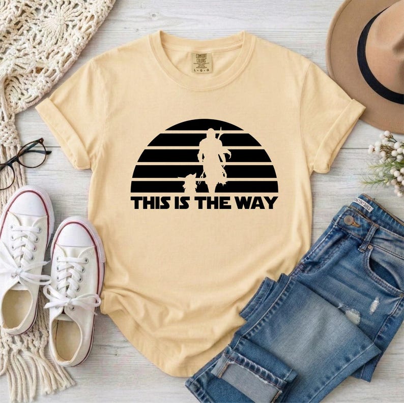 May include: Cream-colored t-shirt featuring a black graphic of a figure and a small companion against a striped background. The text "THIS IS THE WAY" is below the graphic. The shirt is paired with blue jeans and white sneakers.