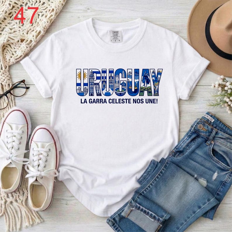 Puede incluir: Una camiseta blanca de manga corta con el texto URUGUAY en letras azules grandes, donde cada letra contiene peque&ntilde;as ilustraciones de monumentos y s&iacute;mbolos. Debajo de la palabra, el texto dice LA GARRA CELESTE NOS UNE!