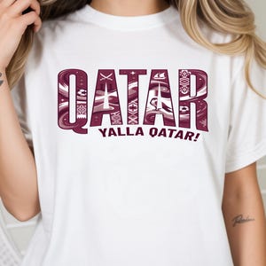 May include: White t-shirt with the word "QATAR" in maroon, featuring intricate patterns within the letters. Below, the text "YALLA QATAR!" is displayed. The shirt is a casual wear item.
