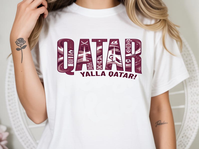 May include: White t-shirt with the word "QATAR" in maroon, featuring intricate patterns within the letters. Below, the text "YALLA QATAR!" is displayed. The shirt is a casual wear item.