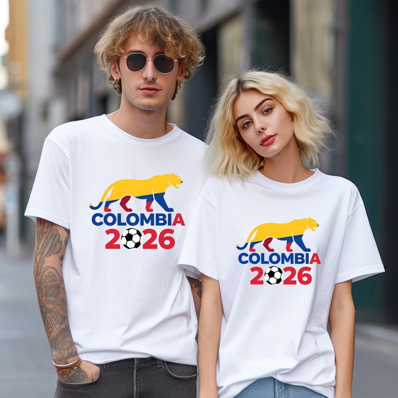 May include: White t-shirts with a colorful graphic featuring a yellow and blue jaguar, the word "COLOMBIA", the number "2026", and a soccer ball. The shirts are worn by two people.