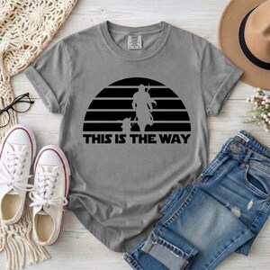 May include: Gray t-shirt featuring a black graphic of a figure and child against a striped sun design, with the text "THIS IS THE WAY" below. The shirt is paired with blue jeans and white sneakers.