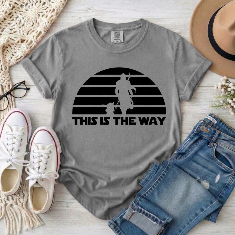 May include: Gray t-shirt featuring a black graphic of a figure and child against a striped sun design, with the text "THIS IS THE WAY" below. The shirt is paired with blue jeans and white sneakers.