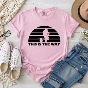 May include: A light pink t-shirt featuring a black graphic of a figure and a child-like character set against a striped sun design. The text "THIS IS THE WAY" is printed below the graphic. The shirt is paired with white sneakers and blue jeans.