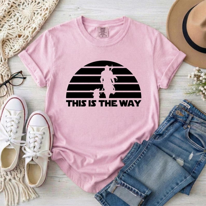 May include: A light pink t-shirt featuring a black graphic of a figure and a child-like character set against a striped sun design. The text "THIS IS THE WAY" is printed below the graphic. The shirt is paired with white sneakers and blue jeans.