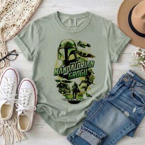 May include: Sage green t-shirt featuring a graphic from The Mandalorian and Grogu. The design includes a helmet, a character, and the text "THIS IS THE WAY". The shirt is paired with blue jeans and white sneakers.