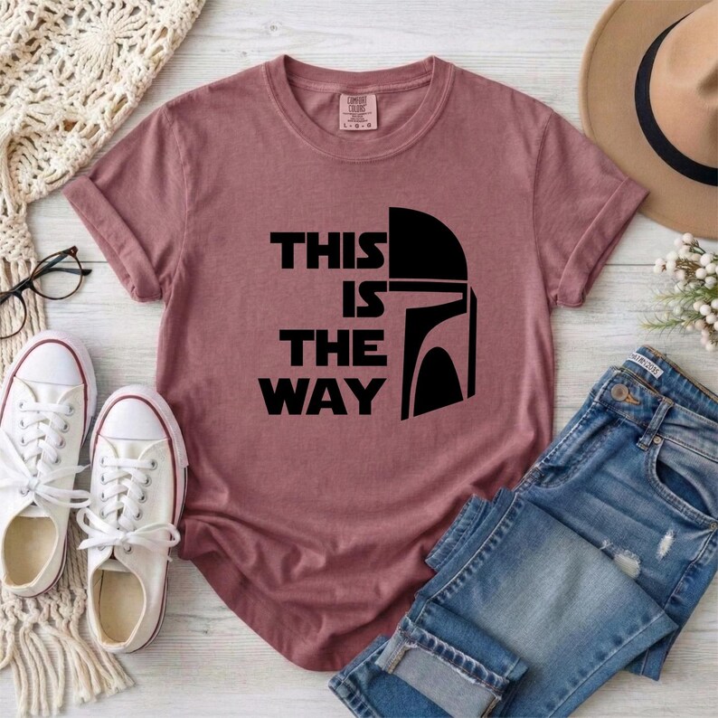 May include: A dusty rose t-shirt with the phrase "THIS IS THE WAY" in black block letters and a black helmet graphic. The shirt is paired with blue jeans, white sneakers, and a tan hat.