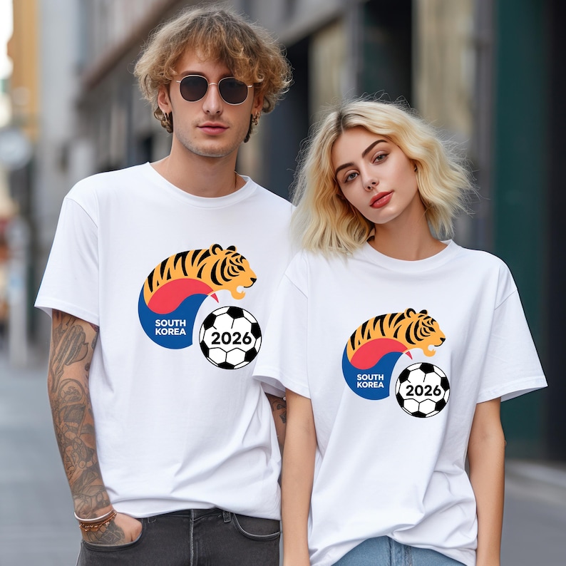 Pu&ograve; includere: T-shirt bianche con un disegno di tigre a strisce nere e dettagli arancioni e gialli, un pallone da calcio e la scritta "SOUTH KOREA 2026". Le magliette sono indossate da due persone.