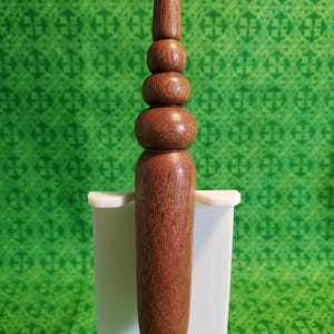 May include: A brown wooden crochet hook handle with a smooth, turned finish. The handle features a series of stacked, rounded decorative segments that taper toward the top, providing a textured grip for the hand while crafting with yarn.