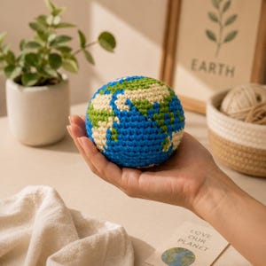 May include: A crocheted globe in blue, green, and cream, held in a hand. The globe features continents and oceans, with the text "LOVE OUR PLANET" visible in the background. The globe is a handmade item.