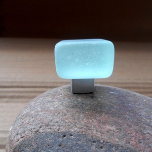 May include: A light blue square glass knob with a white base, sitting on a textured rock.