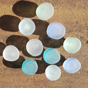 May include: A collection of ten smooth, round glass stones in various shades of blue, green, and white. The stones are arranged in a triangular pattern on a brown, textured surface.