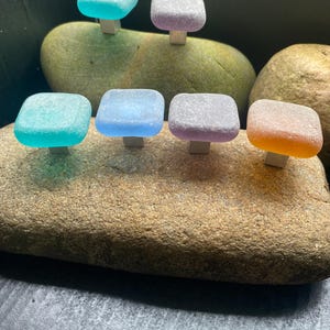 May include: A collection of six colorful, square-shaped cabinet knobs. The knobs are in shades of blue, purple, and orange, mounted on small, silver-colored metal bases. They are displayed on a large, textured, brown rock, with a dark background.