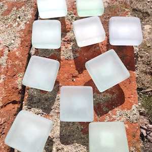 May include: Ten frosted glass squares in various shades of white and light blue, arranged in a pattern on a red brick surface.