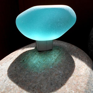 May include: A smooth, translucent, light blue sea glass piece with a frosted texture, attached to a small, square, silver-toned metal base. The object is positioned atop a large, rounded, speckled tan stone, casting a distinct shadow.
