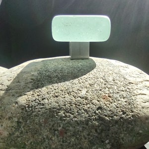 May include: A light green glass bottle stopper with a silver metal base, sitting on a gray, textured rock.