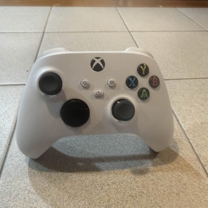 May include: A white Xbox controller with black thumbsticks and colored action buttons. The controller has the Xbox logo and various function buttons. The controller is resting on a tiled surface.