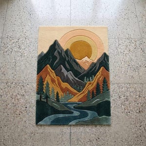May include: A rectangular rug featuring a mountain landscape design. The rug depicts a river winding through a valley, with mountains in shades of green, gray, and orange. A yellow sun and a light beige sky complete the scene.