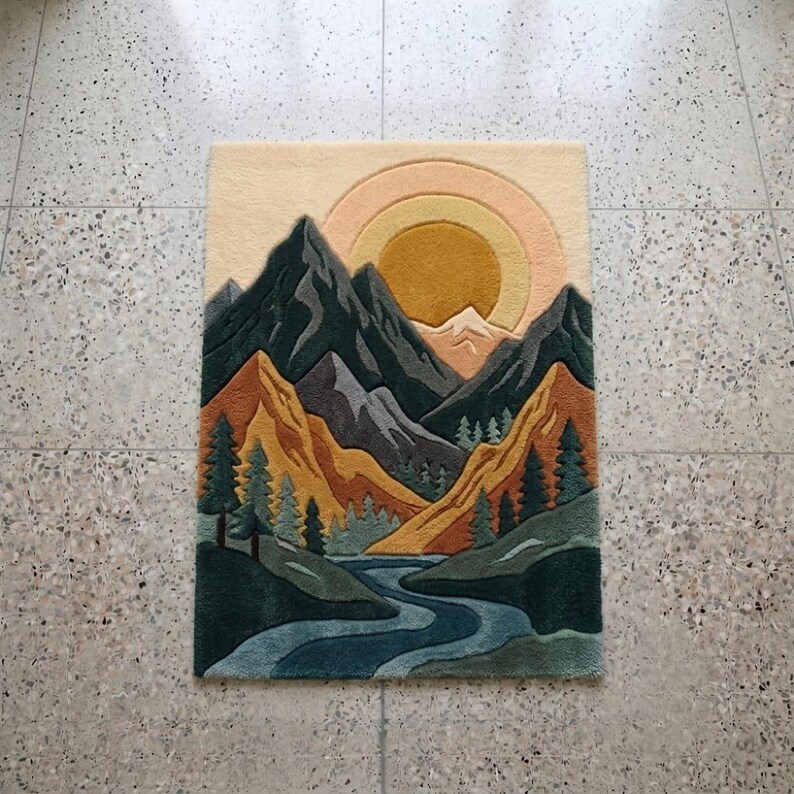 May include: A rectangular rug featuring a mountain landscape design. The rug depicts a river winding through a valley, with mountains in shades of green, gray, and orange. A yellow sun and a light beige sky complete the scene.
