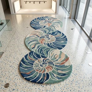 May include: Three round rugs with a nautilus shell design in shades of blue, green, and beige are arranged in a row on a speckled floor. The rugs have a spiral pattern and are placed in a well-lit, modern space.