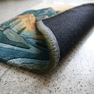 May include: A rolled-up rug with a landscape design featuring mountains, trees, and water in shades of blue, green, and yellow. The rug has a dark gray backing and a green stitched border.