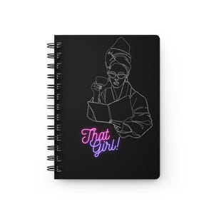 May include: Black spiral-bound notebook featuring a line drawing of a person in a robe, reading and drinking. The text "That Girl!" is in pink and purple neon-style lettering.