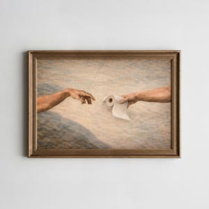 May include: A framed print parodying Michelangelo's 'The Creation of Adam'. The image shows two arms reaching out, with one offering a roll of toilet paper to the other. The frame is a warm, golden brown, adding a classic touch to the humorous artwork.