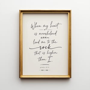 May include: A framed print with a gold-colored frame. The print features elegant black script that reads "When my heart is overwhelmed, lead me to the rock that is higher than I." The text is from Psalm 61:2.
