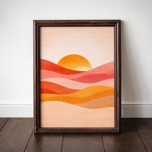 Könnte beinhalten: Ein gerahmter Kunstdruck mit einer minimalistischen Landschaft mit Sonnenuntergang. Das Kunstwerk zeigt geschichtete, wellenförmige Hügel in Orange-, Rot- und Pfirsichtönen mit einer leuchtend orangefarbenen Sonne. Der Rahmen ist dunkelbraun.