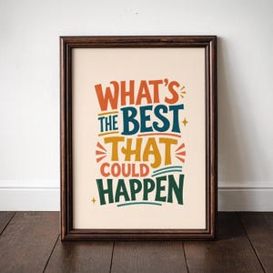 May include: Framed art print with the text "WHAT'S THE BEST THAT COULD HAPPEN" in colorful, stylized lettering. The print is in a dark brown wooden frame, set against a cream-colored background. The artwork is displayed on a wooden floor.