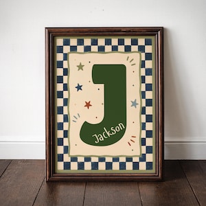 May include: A framed wall art print featuring a large dark green letter J with the name Jackson written in white script. The central cream-coloured panel is decorated with small multi-coloured stars and dots, surrounded by a navy blue and white checkerboard border.