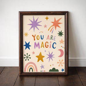 May include: A framed art print with a cream background and colorful illustrations of stars, a rainbow, and a crescent moon. The words "YOU ARE MAGIC" are written in various colors. The frame is a dark brown color.