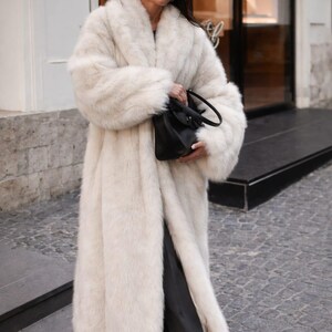 May include: A long, ivory-colored faux fur coat with a wide collar and sleeves. The coat is worn with dark pants and a black handbag. The person is wearing sunglasses and standing on a cobblestone street.
