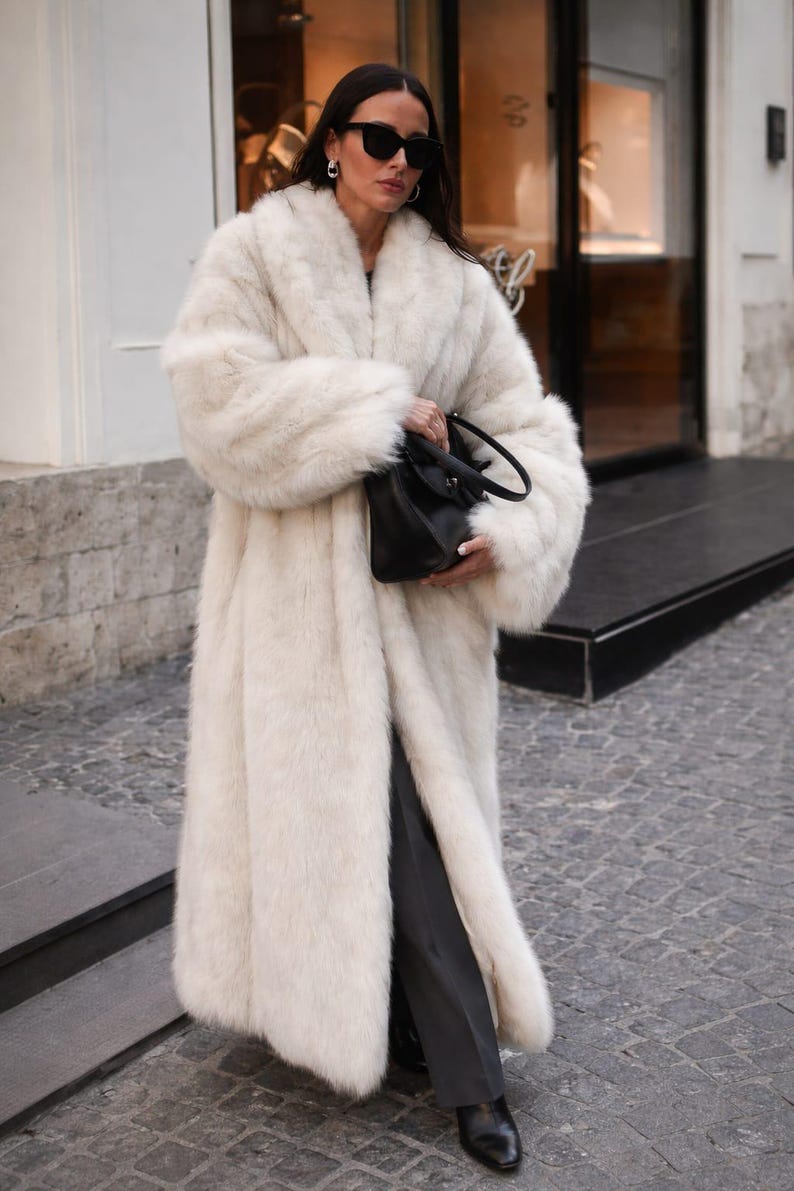 May include: A long, ivory-colored faux fur coat with a wide collar and sleeves. The coat is worn with dark pants and a black handbag. The person is wearing sunglasses and standing on a cobblestone street.