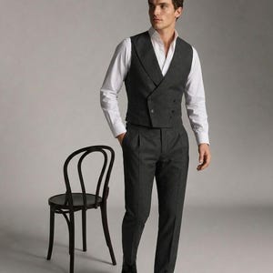 May include: A charcoal gray three-piece suit with a double-breasted vest, white dress shirt, and matching trousers. The suit is paired with black dress shoes. A black wooden chair is to the left of the subject.