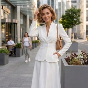May include: A woman wearing a white suit with a V-neck jacket, a fitted waist, and a long skirt. She carries a small brown purse and smiles. The background includes a city street and a building with a "BOUTIQUE" sign.