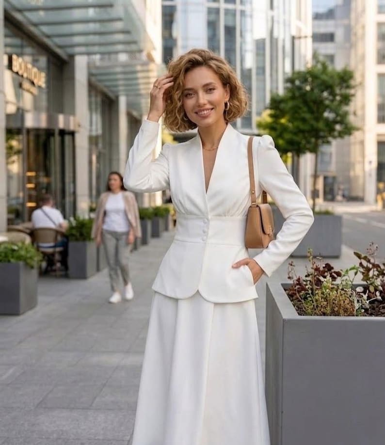 May include: A woman wearing a white suit with a V-neck jacket, a fitted waist, and a long skirt. She carries a small brown purse and smiles. The background includes a city street and a building with a "BOUTIQUE" sign.