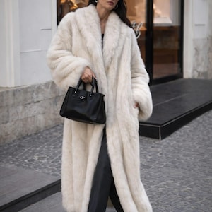 May include: A long, cream-colored faux fur coat with a wide collar. The coat is worn with black pants, black shoes, and a black handbag. The person is wearing sunglasses and silver hoop earrings. The coat appears to be floor-length.