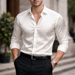 Puede incluir: Camisa blanca de manga larga, pantalones negros y cinturón negro. Se ve un reloj de pulsera plateado en la muñeca izquierda. Adecuado para ocasiones formales o de negocios.