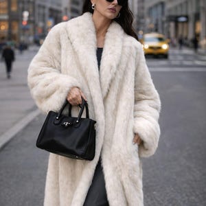 May include: A woman wearing a long, cream-colored faux fur coat and black sunglasses, holding a black handbag. The coat has a wide collar and a soft, textured appearance. The background shows a city street with buildings and a yellow taxi.