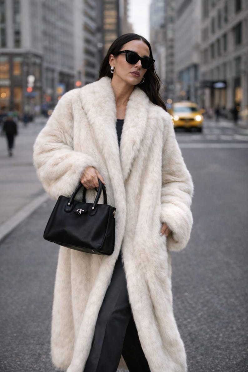 May include: A woman wearing a long, cream-colored faux fur coat and black sunglasses, holding a black handbag. The coat has a wide collar and a soft, textured appearance. The background shows a city street with buildings and a yellow taxi.