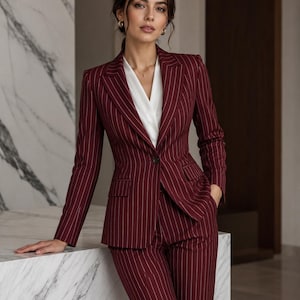 May include: A dark burgundy pinstriped blazer and matching trousers set featuring a single-button closure, notched lapels, and flap pockets, worn over a crisp white V-neck blouse. The tailored suit has a structured fit with long sleeves.