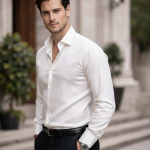 Puede incluir: Un hombre con una camisa blanca abotonada, pantalones negros y un cintur&oacute;n negro. Tambi&eacute;n lleva un reloj. La camisa est&aacute; desabrochada en la parte superior. El fondo es un edificio con columnas.