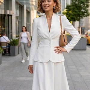 May include: A woman in a white suit, consisting of a blazer and a midi skirt, stands outdoors. The blazer has a V-neck and button closure. She carries a small brown purse. The background includes a building and other people.
