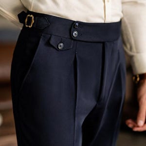 May include: Close-up of navy blue dress pants with a high waist and pleats. The pants feature a black belt with a gold buckle and button details. The pants are part of a formal wear ensemble.