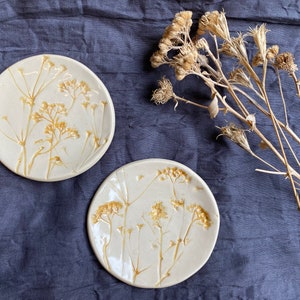 May include: Two white ceramic plates with a pressed flower design. The plates have a subtle yellow hue and feature delicate pressed flowers in a light brown color.